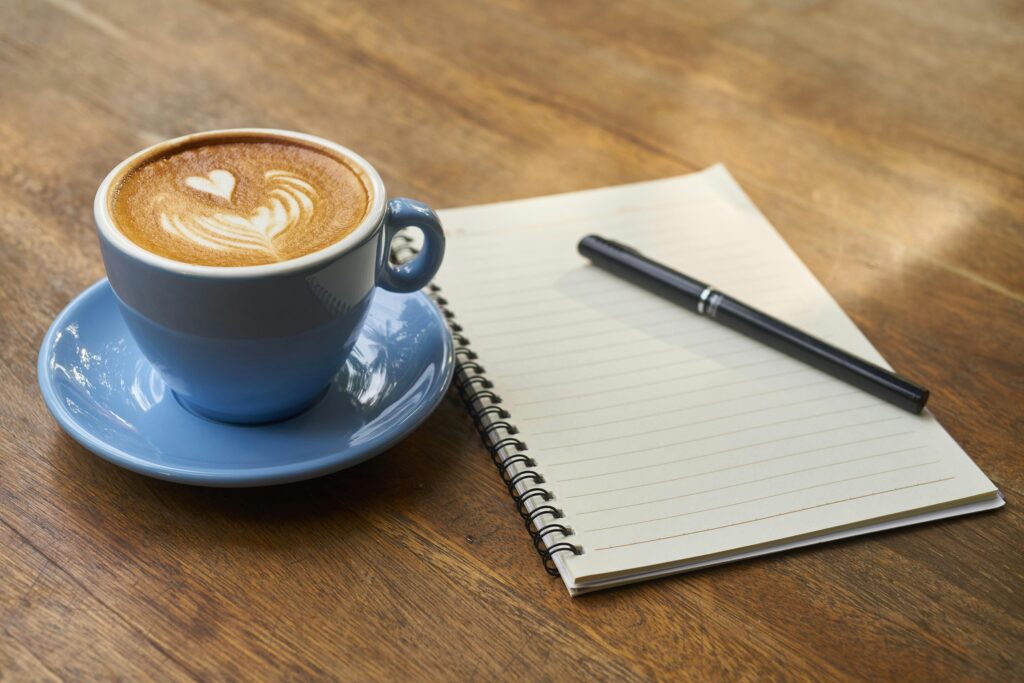 A calming scene of a latte with art in a blue cup beside a notebook and pen on a wooden table.